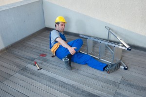 A man wearing a hard hat is lying on the ground next to a fallen ladder while holding his leg